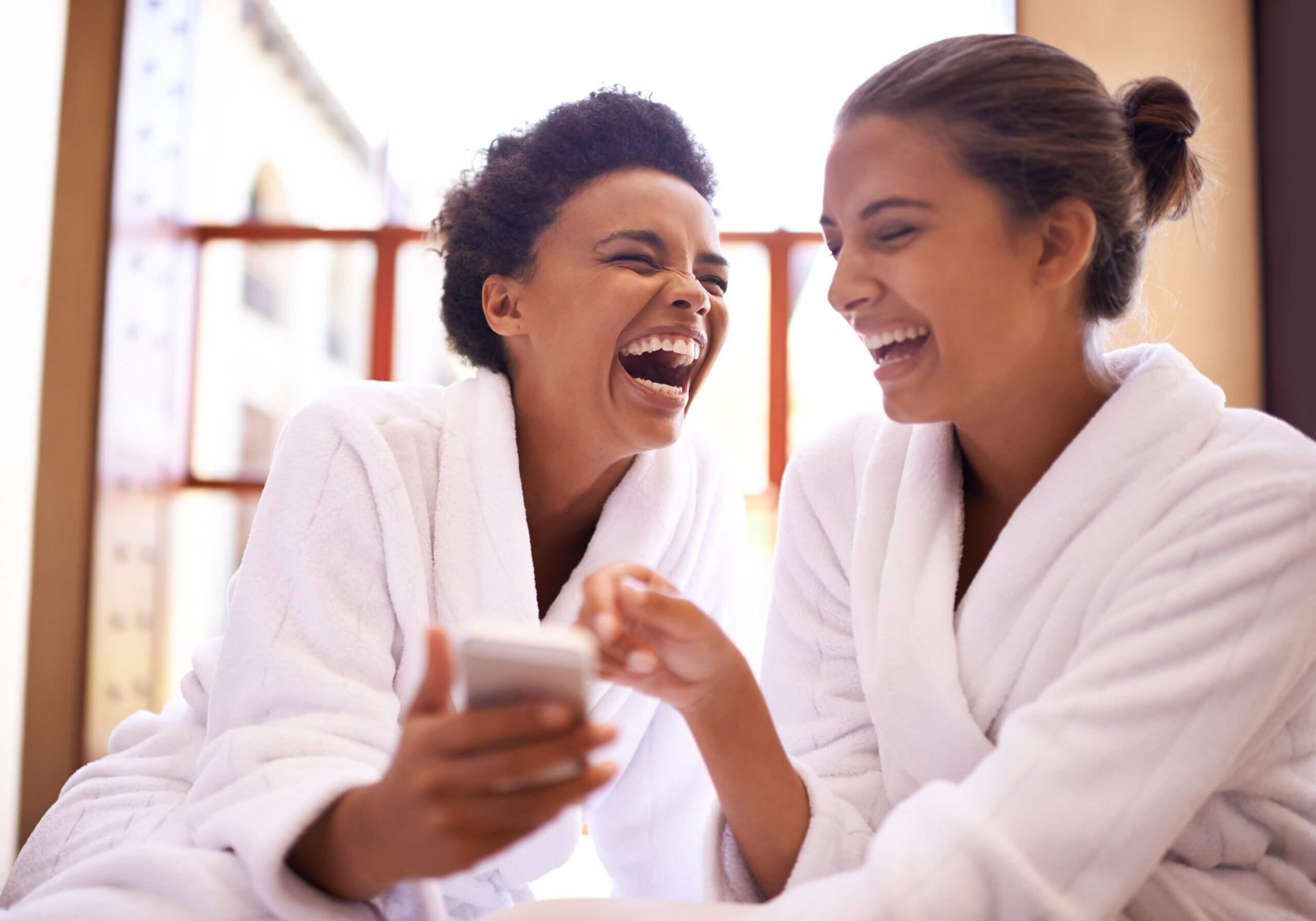 Beauty, phone and spa with women laughing in robes for luxury pampering or treatment together. Happy, young friends at resort or salon for wellness or weekend getaway