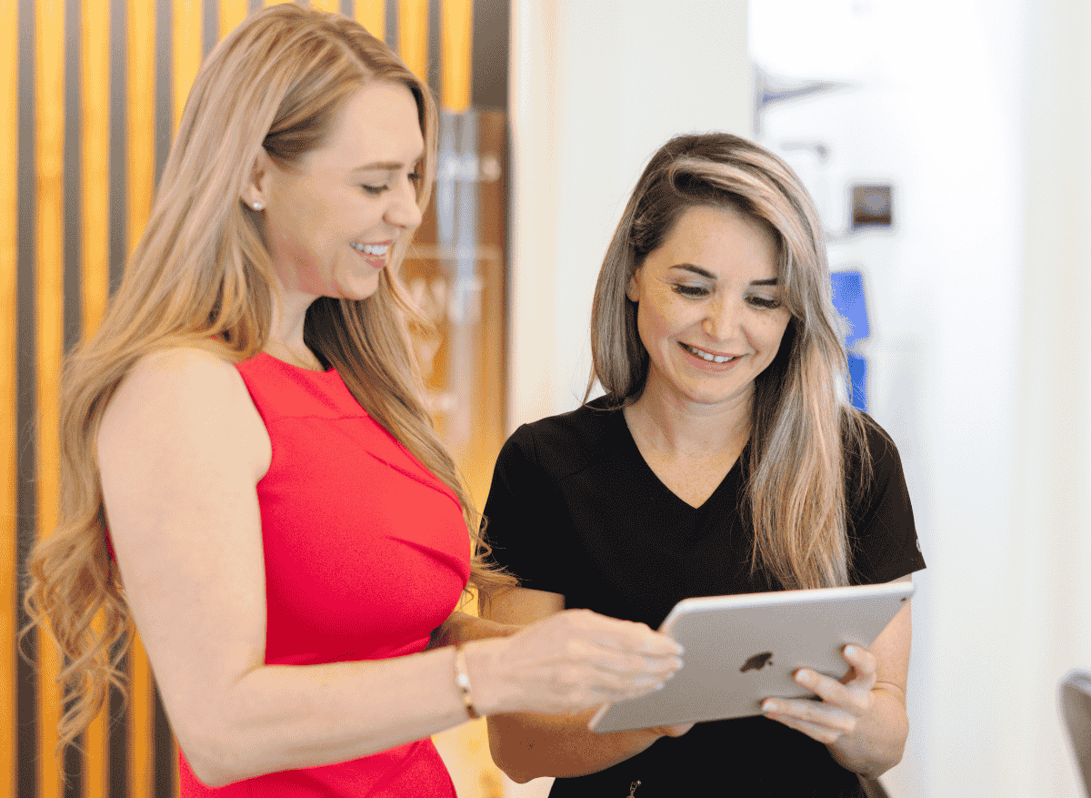 Two women standing indoors, smiling and looking at a tablet together.