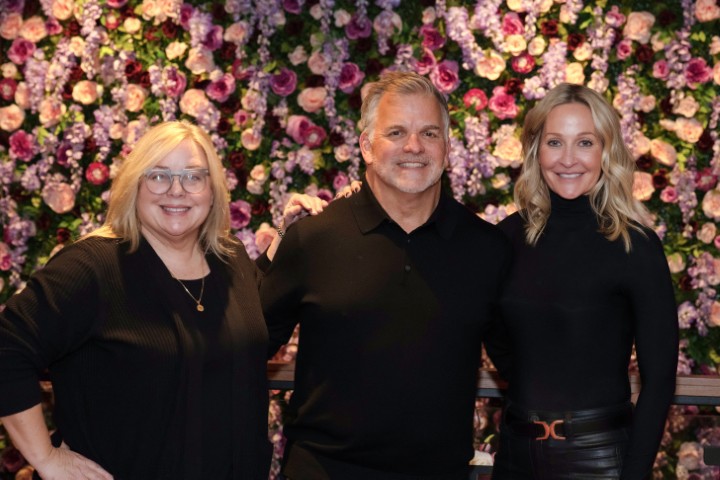 Three adults stand together in front of a wall covered with purple and pink flowers, all wearing black clothing and smiling at the camera.